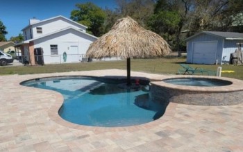 Custom backyard swimming pool with stone patio and thatched umbrella beside a house